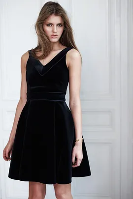 Woman in a sleeveless black velvet dress stands against a white paneled wall.