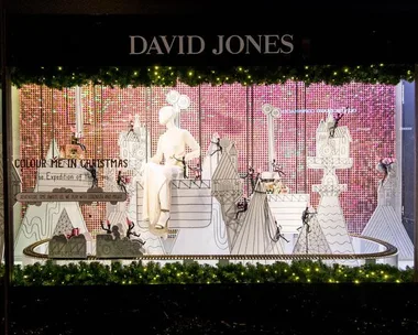 David Jones Christmas window display featuring a mannequin, festive decorations, and a miniature Christmas village with lights and greenery.