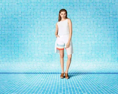 Woman in white sleeveless top and colorful shorts posed against a blue tiled background.