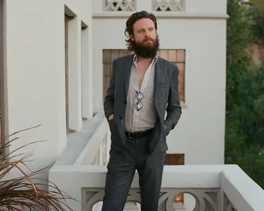 Father John Misty standing on a balcony, wearing a suit with an unbuttoned shirt and sunglasses hanging on his chest.