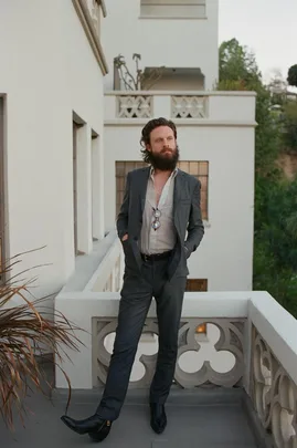 Father John Misty standing on a balcony, wearing a suit with an unbuttoned shirt and sunglasses hanging on his chest.