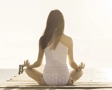 A woman in white clothing sits cross-legged on a wooden pier, meditating by the ocean under a bright sky.