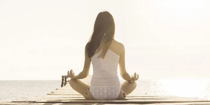 A woman in white clothing sits cross-legged on a wooden pier, meditating by the ocean under a bright sky.