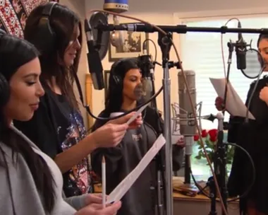 Kim Kardashian and her sisters making a surprise video tribute for Kris Jenner, standing with microphones and reading scripts.