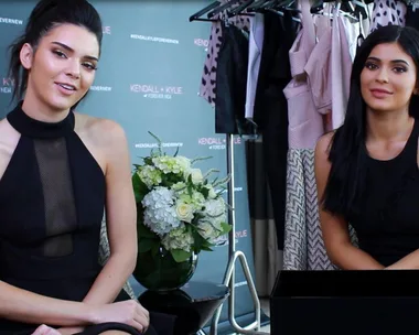 Kendall and Kylie Jenner sitting together in black outfits during an interview with a clothing rack in the background.