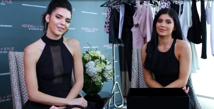 Kendall and Kylie Jenner sitting together in black outfits during an interview with a clothing rack in the background.