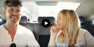 Two people smiling and talking while sitting in the backseat of a car on a sunny day, part of a video from "elle-tvs-beauty-cab."