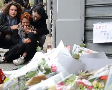 Three people mourning near a large collection of flowers and notes with a visible sign that says "LOVE over Fear & Hate."