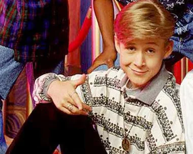 Young actor sitting with a smile, wearing a patterned shirt; part of a group photo in a teen drama scene.