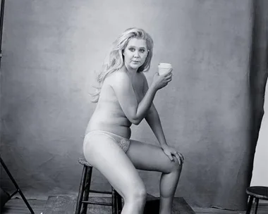 A woman in underwear and heels poses sitting on a stool, holding a coffee cup, against a plain backdrop.