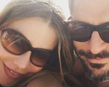 Couple wearing sunglasses smiles while lying on a beach towel, sunlit and relaxed.