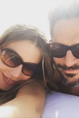 Couple wearing sunglasses smiles while lying on a beach towel, sunlit and relaxed.