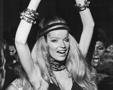 Person with long hair in a black bikini dancing at a party, wearing bracelets and a headband, arms raised in joy.