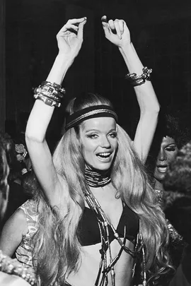 Person with long hair in a black bikini dancing at a party, wearing bracelets and a headband, arms raised in joy.