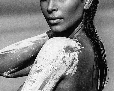 Black-and-white image of a woman with wet hair, partially covered in white paint, posing in a desert setting.