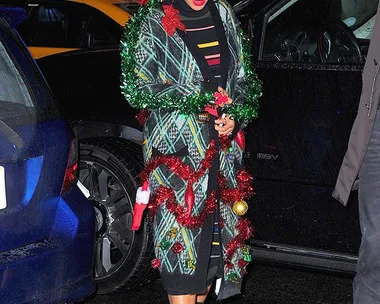 Person in festive plaid sweater, Santa hat, and Christmas decorations, standing by cars at night.