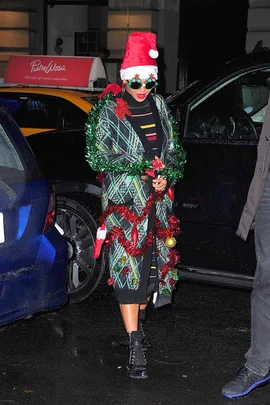 Person in festive plaid sweater, Santa hat, and Christmas decorations, standing by cars at night.