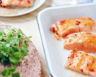 Baked salmon with sweet chili sauce served in a white baking dish alongside a plate with garnished salmon and a herb garnish.