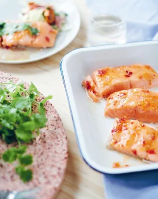 Baked salmon with sweet chili sauce served in a white baking dish alongside a plate with garnished salmon and a herb garnish.