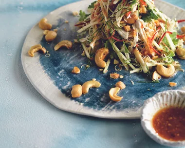 Shredded chicken salad with herbs, cashews, and thinly sliced vegetables, served with a side dish of dipping sauce.