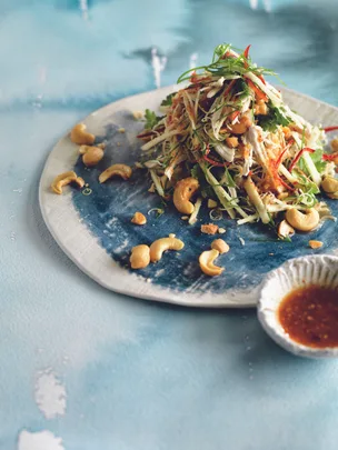 Shredded chicken salad with herbs, cashews, and thinly sliced vegetables, served with a side dish of dipping sauce.