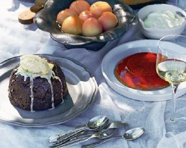 A table setting featuring Christmas pudding with cream, a flan, fresh peaches, whipped cream, and a glass of white wine.