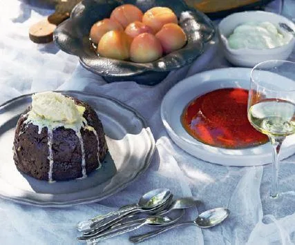 A table setting featuring Christmas pudding with cream, a flan, fresh peaches, whipped cream, and a glass of white wine.