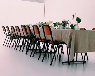 Long table with a beige tablecloth, surrounded by orange chairs, and decorated with greenery and small floral arrangements.