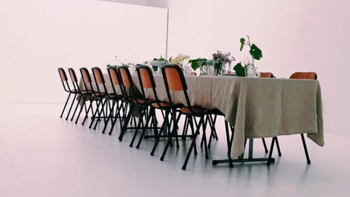 Long table with a beige tablecloth, surrounded by orange chairs, and decorated with greenery and small floral arrangements.