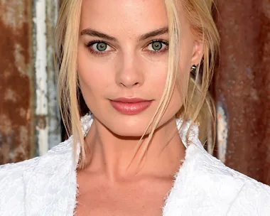 A close-up image of a woman with blonde hair and green eyes, wearing a white garment. She is standing against a textured background.