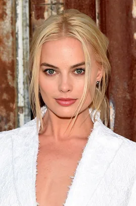 A close-up image of a woman with blonde hair and green eyes, wearing a white garment. She is standing against a textured background.