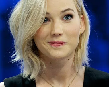 Jennifer Lawrence with short blonde hair, wearing a black dress and a star-shaped necklace, looking amused.