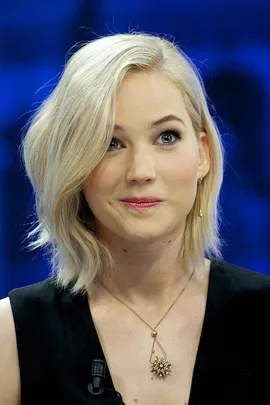 Jennifer Lawrence with short blonde hair, wearing a black dress and a star-shaped necklace, looking amused.