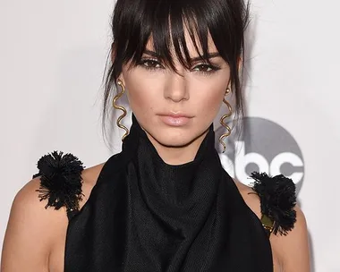 Woman with dark hair in a high bun, wearing a black sleeveless outfit with decorative details on the shoulders, at an event.