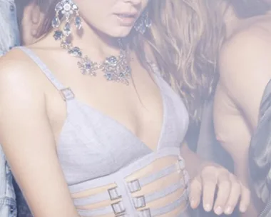 A woman in a light-colored dress with strap details and statement jewelry at a crowded, smoky party.