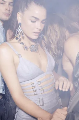 A woman in a light-colored dress with strap details and statement jewelry at a crowded, smoky party.