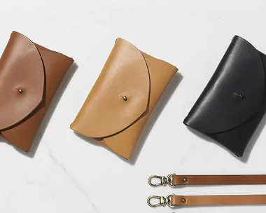 Three leather clutch bags in brown, tan, and black, featuring snap closures and two detachable leather straps with metal clasps.