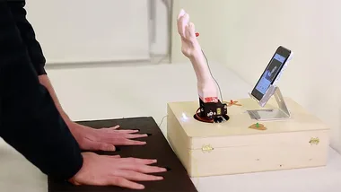 A person placing their hands on black sensors connected to a mechanical hand swiping on a smartphone dating app.