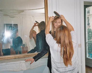 A hairstylist fixes a model's hair in front of a large mirror in a bright, elegant room during a photoshoot.