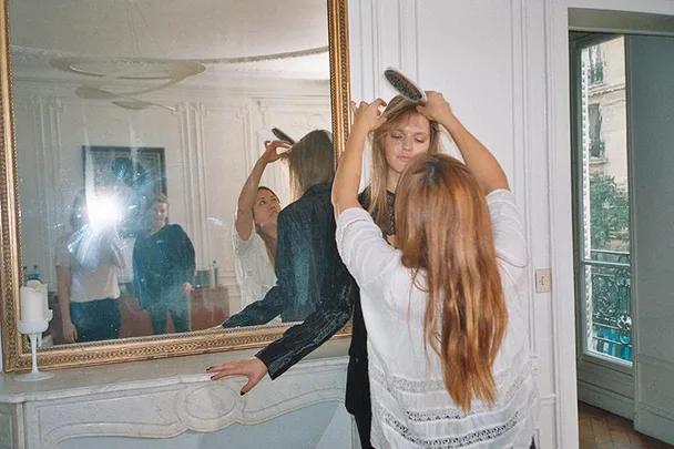 A hairstylist fixes a model's hair in front of a large mirror in a bright, elegant room during a photoshoot.