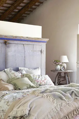 Rustic bedroom with a wooden headboard, floral bedding, and a side table with a lamp and vase. Cozy and inviting atmosphere.