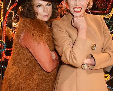 Two women, one in a brown fur vest and hat, the other in a beige coat, posing in front of colorful neon lights.