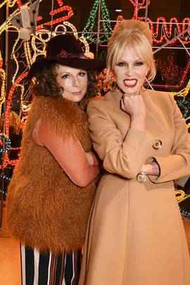 Two women, one in a brown fur vest and hat, the other in a beige coat, posing in front of colorful neon lights.