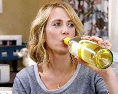 Woman in a gray shirt drinking white wine straight from the bottle in a cozy living room.