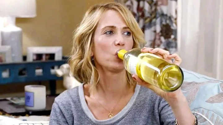 Woman in a gray shirt drinking white wine straight from the bottle in a cozy living room.