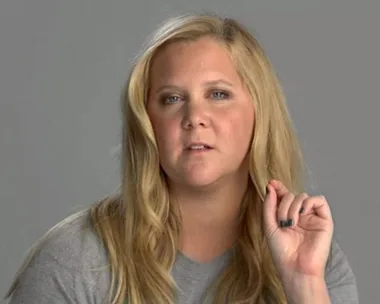 Person with long blonde hair dramatically recites lines, wearing a grey shirt with a neutral background.