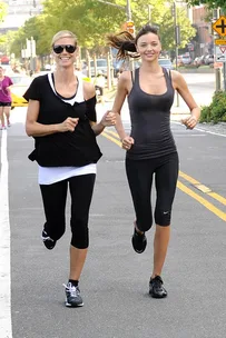 Two women jogging on a city street, wearing athletic clothing and sneakers, with buildings and trees in the background.