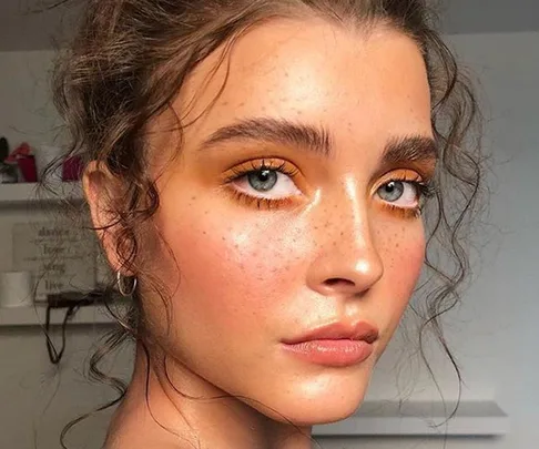 Young woman with curly hair and fake freckles, wearing orange eyeshadow, hoop earrings, and a neutral lip color.