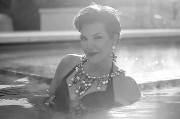 "Kris Jenner in a black swimsuit and statement jewelry, emerging from a pool in a black and white photo for Love Advent."