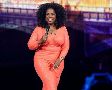 Oprah Winfrey in a coral dress holding a microphone on stage with a bridge backdrop.